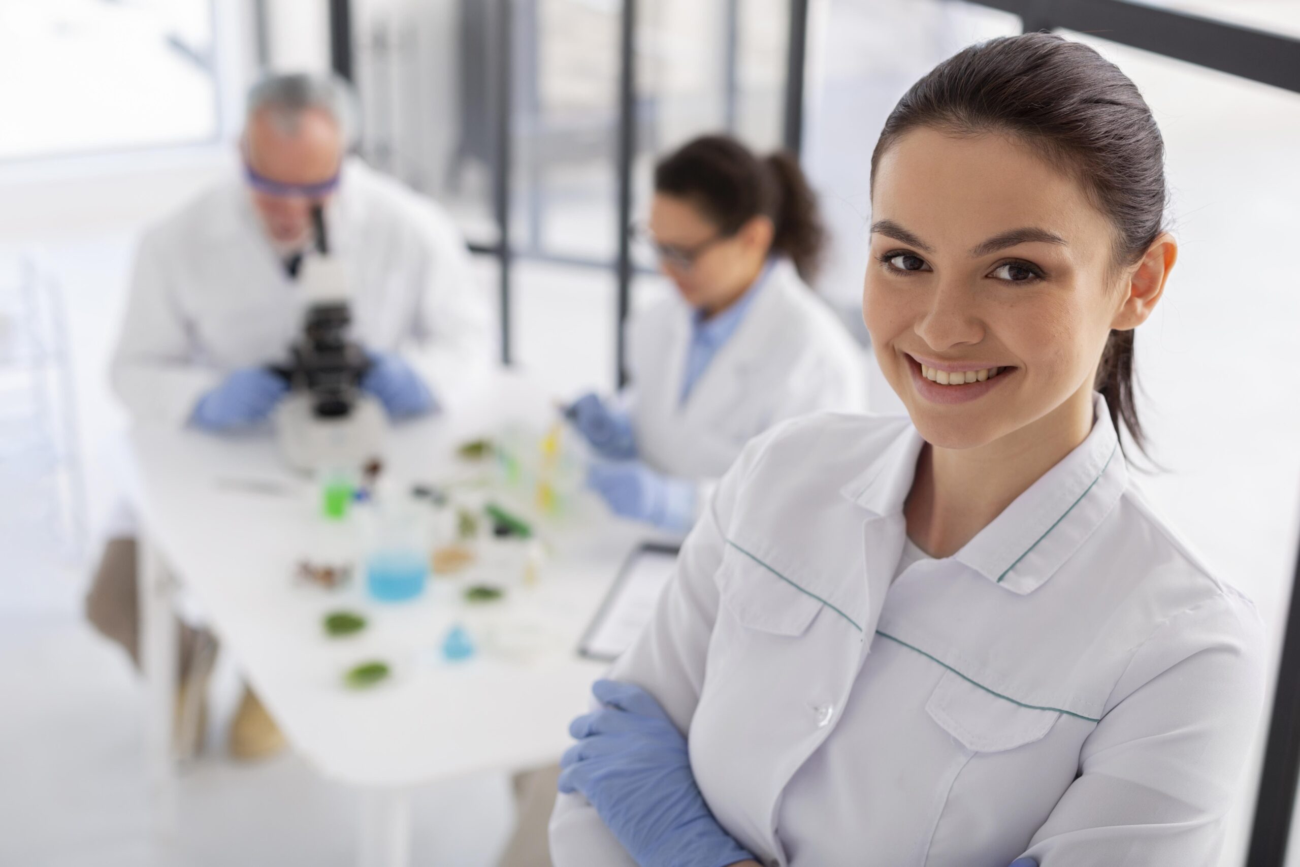 Inicio Profesional de laboratorio clínico sonriendo en un entorno moderno con su equipo al fondo realizando análisis científicos.